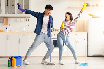 Funny asian loving couple singing songs while cleaning kitchen, using broom and mop as microphones,...