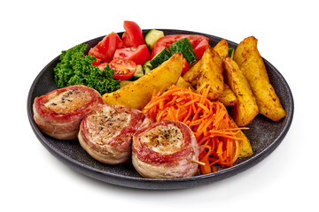 Grilled bacon-wrapped pork medallions with potato wedges, carrot salad, and fresh vegetables on a black plate, isolated on white background.