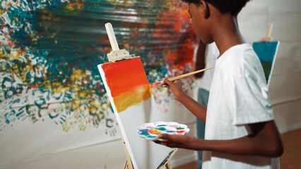 Closeup of african boy hand paint canvas with warm tone color while caucasian girl in casual outfit...