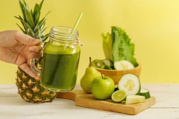 Hand holding green smoothie in mason jar with pineapple, apple, cucumber, lime, and lettuce on a wooden table, yellow background.