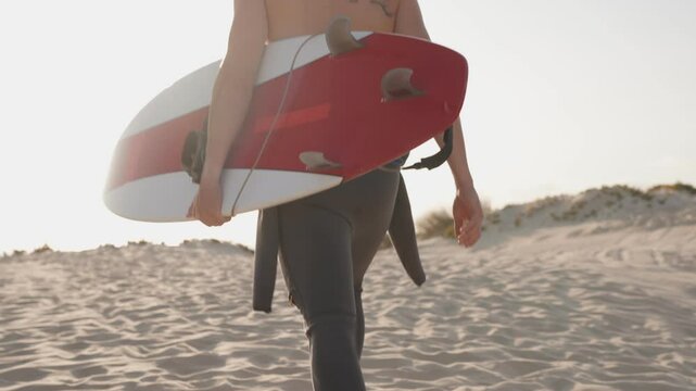A surfer walks across a sandy beach carrying their surfboard after a surf session at sunset. Active lifestyle pursuit.