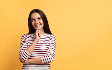 Positive young brunette woman looking at copy space, touching chin and smiling, isolated on yellow studio background