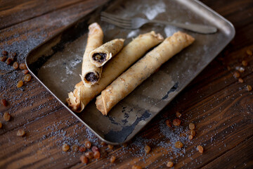 Golden-brown rolled pancakes with a rich poppy seed and raisin filling displayed on a vintage metal tray. The wooden surface is dusted with poppy seeds and raisins for a rustic, appetizing presentatio