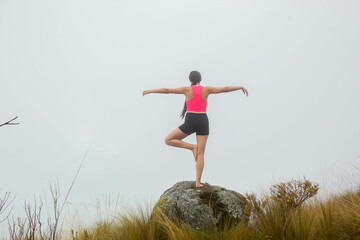 Joven atractiva practicando yoga al aire libre rodeada de naturaleza y nubes, simbolizando un estilo de vida saludable y equilibrio interior