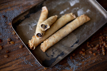 Golden-brown rolled pancakes with a rich poppy seed and raisin filling displayed on a vintage metal tray. The wooden surface is dusted with poppy seeds and raisins for a rustic, appetizing presentatio