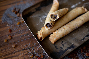 Rolled pancakes with sweet prune filling and poppy seed topping, served on a rustic baking tray, surrounded by raisins and powdered sugar on a wooden table.