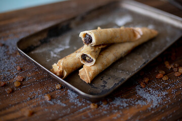 Rolled pancakes with sweet prune filling and poppy seed topping, served on a rustic baking tray, surrounded by raisins and powdered sugar on a wooden table.