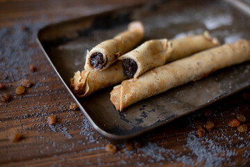 Rolled pancakes with sweet prune filling and poppy seed topping, served on a rustic baking tray, surrounded by raisins and powdered sugar on a wooden table.