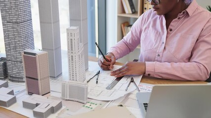 African woman architect focused on drawing city blueprint with scale models. Professional workspace includes laptop and drafting tools, highlighting creativity and precision in architectural design.