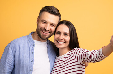 A happy European couple smiles brightly while embracing in front of a vibrant yellow background, showcasing their love and affection for one another during a playful moment.