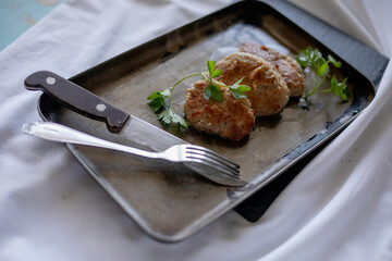 A trio of juicy meat patties is artfully arranged on a vintage baking tray, garnished with fresh parsley leaves. Fork and knife are placed next to the tray on a rustic white cloth, highlighting the ho