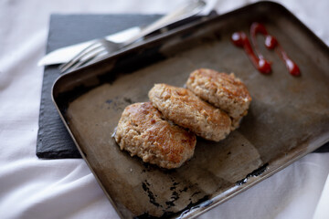 Three golden-brown homemade meat patties are placed on a vintage metal tray. The tray rests on a white tablecloth, with cutlery in the background, adding a rustic aesthetic to the composition.