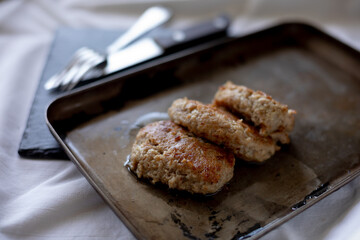 Freshly fried meat patties rest on a slightly worn metal tray, with visible oil drops enhancing their golden appearance. The cutlery peeks from the background on a dark surface, giving a rustic, homel