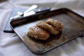 Freshly fried meat patties rest on a slightly worn metal tray, with visible oil drops enhancing their golden appearance. The cutlery peeks from the background on a dark surface, giving a rustic, homel