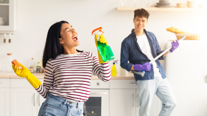 Joyful asian woman singing songs and dancing with closed eyes while cleaning kitchen at home,...