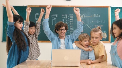 Teacher and multicultural student looking at laptop while celebrate success project and put hands in the air. Group of children smiling to camera while standing at blackboard in classroom. Pedagogy.