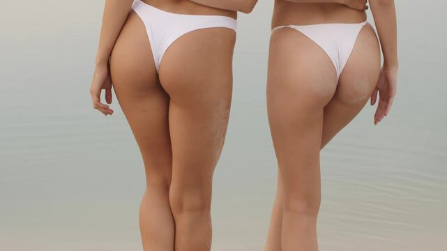 Two women in matching white bikinis stand together near calm water, embracing and raising their arms in joy. The serene scene radiates themes of friendship, love, and carefree summer moments.