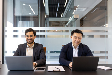Two professionals are seated at a table, each using a laptop for work. They appear engaged and...
