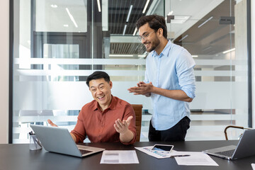 Fototapeta premium Two coworkers are seen in bright, modern office setting, energetically discussing and celebrating work success. The table displays graphs and a smartphone, emphasizing a productive workplace ambiance
