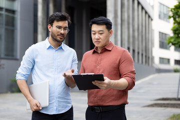 Two professionals engage in a discussion outdoors, reviewing information on a clipboard. One holds...