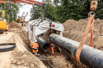 Zwei Bauarbeiter leiten den Einsatz eines Krans an einer Baustelle, um große Rohre in einem tiefen...