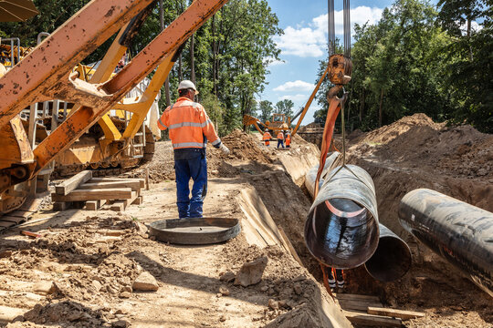 Auf der Baustelle werden Rohre mit Hilfe eines Krans verlegt. Bauarbeiter beaufsichtigen die Montage und Positionierung der gro&szlig;en Rohre in einem tiefen Graben