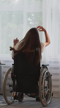 Lonely lady with disability with multiple sclerosis looks out bright window in room. Brunette woman sits in wheelchair touching long loose hair backside view