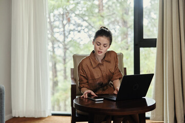 Young woman working on a laptop in a cozy indoor setting, showcasing a stylish brown outfit, focused on productivity amidst a serene natural backdrop