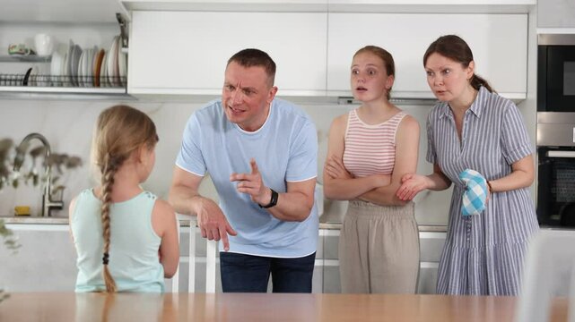 At home kitchen, parents scold daughter for careless act. Sulky preschool girl stand near table during serious conversation with parents