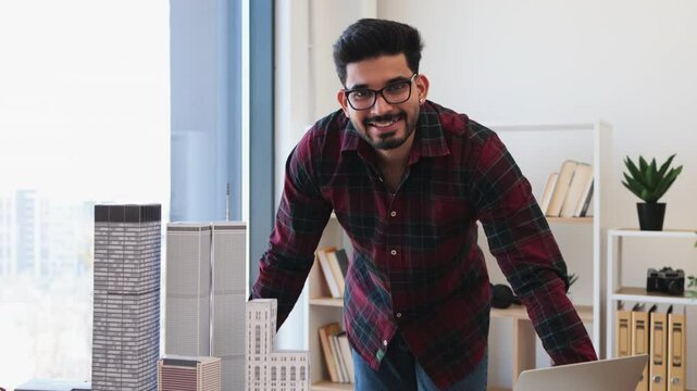 Indian male architect in 30s designing city architecture model at office desk with laptop and buildings. Creative professional working on urban development project in modern workspace.
