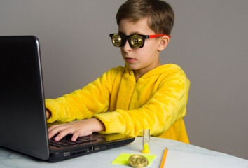 Boy in yellow pajamas and wearing glasses with bitcoin sits at the computer, earning on Bitcoin mining. The child learns to make money.