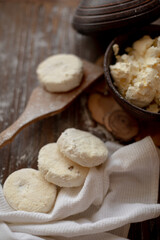 A rustic wooden table with cottage cheese pancakes (syrniki) on a wooden spoon, accompanied by a bowl of fresh cottage cheese. The flour-dusted surface creates a cozy and warm atmosphere