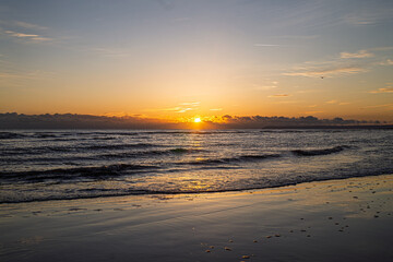 Fototapeta premium West St Leonards beach at sunset. The scene shows the beach at low ride, exposing the sand. Waves roll into the beach. The setting sun creates contrasting colours and deep shadows.