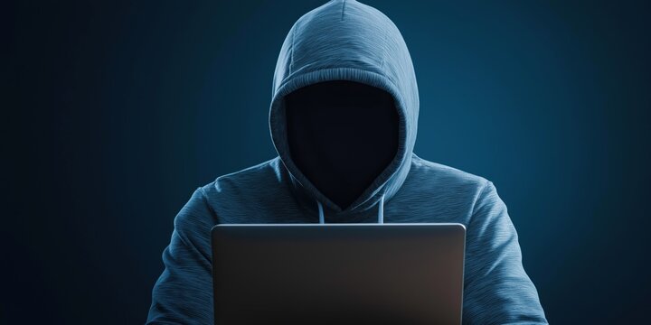 A hooded individual using a laptop symbolizing anonymity, cybersecurity, and the digital age in dim lighting.