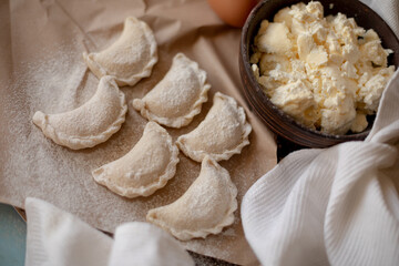 Fresh dumplings (varenyky) filled with cottage cheese, arranged on parchment paper alongside a bowl of cottage cheese and a white kitchen towel. The wooden background and flour-dusted details 