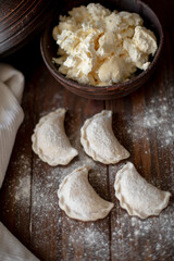 Fresh dumplings (varenyky) filled with cottage cheese, arranged on parchment paper alongside a bowl of cottage cheese and a white kitchen towel. The wooden background and flour-dusted details 
