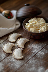 Fresh dumplings (varenyky) filled with cottage cheese, arranged on parchment paper alongside a bowl of cottage cheese and a white kitchen towel. The wooden background and flour-dusted details 