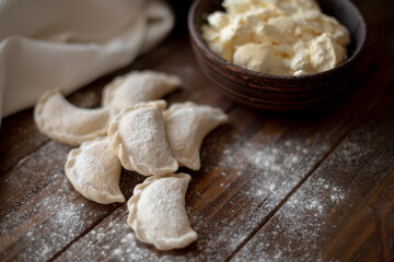 Fresh dumplings (varenyky) filled with cottage cheese, arranged on parchment paper alongside a bowl of cottage cheese and a white kitchen towel. The wooden background and flour-dusted details 