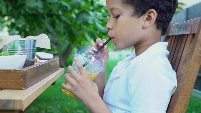 African American teenager drinks lemonade with straw in the park outdoors, curly schoolboy drinks juice from plastic glass in nature