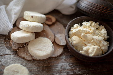 Raw cottage cheese pancakes (syrniki) with raisins, dusted with flour, arranged on a rustic wooden board alongside a dark wooden bowl filled with fresh cottage cheese. A cozy kitchen atmosphere 