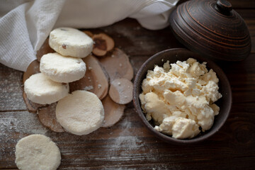 Raw cottage cheese pancakes (syrniki) with raisins, dusted with flour, arranged on a rustic wooden board alongside a dark wooden bowl filled with fresh cottage cheese. A cozy kitchen atmosphere 