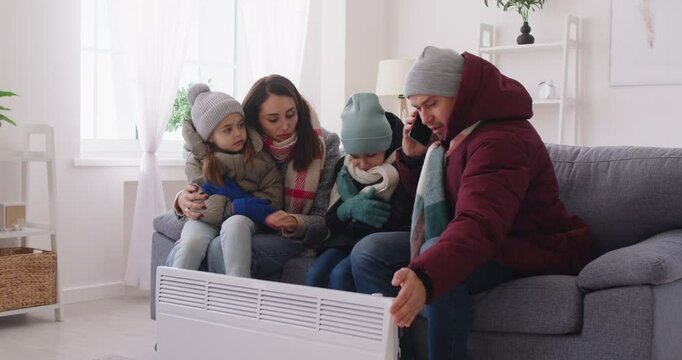 Frozen young family with kids in winter outwear sitting on sofa in living room near electric heater at home warning up calling public utility service by mobile phone. Heating problems. 4k video.
