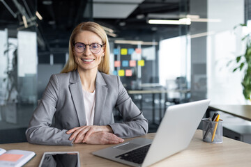 Mature woman businesswoman wearing glasses and suit seated at desk in modern office. Smiling and confident, she works with a laptop and notepad, showcasing professional productivity and success.