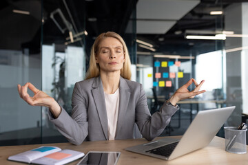 Mature businesswoman seated at her desk performing mindfulness meditation to relax and focus. Maintaining a calm and composed expression, she sits surrounded by work materials