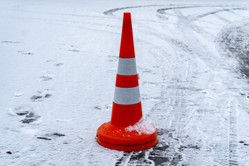 A bright orange traffic cone is placed on a snowy surface in a parking lot, showing tire tracks and footprints around it