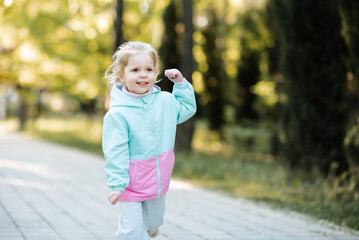 Cute kid girl 4-5 year old wearing sport jacket running outdoor in park. Looking at camera. Childhood. Springtime.