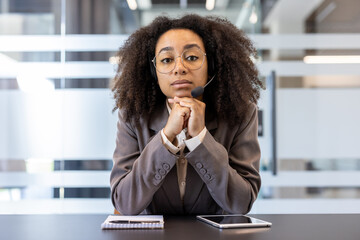 Portrait of a young African American woman in a suit sitting in an office at a desk wearing a headset and with her hands folded together looking seriously at the camera