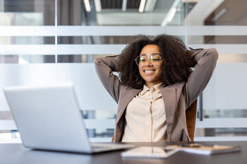 Smiling and satisfied young African-American woman sitting in the office at the desk with her eyes...