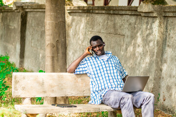 Fototapeta premium Young cheerful black man using mobile phone and laptop computer. Modern lifestyle