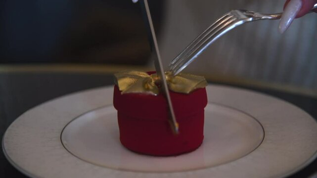 Close-up side view of woman cutting in half red gift box shaped dessert and showing it's filling (brownie,  cherry and cream with chocolate bow) on white table in cafe or restaurant. Handheld video